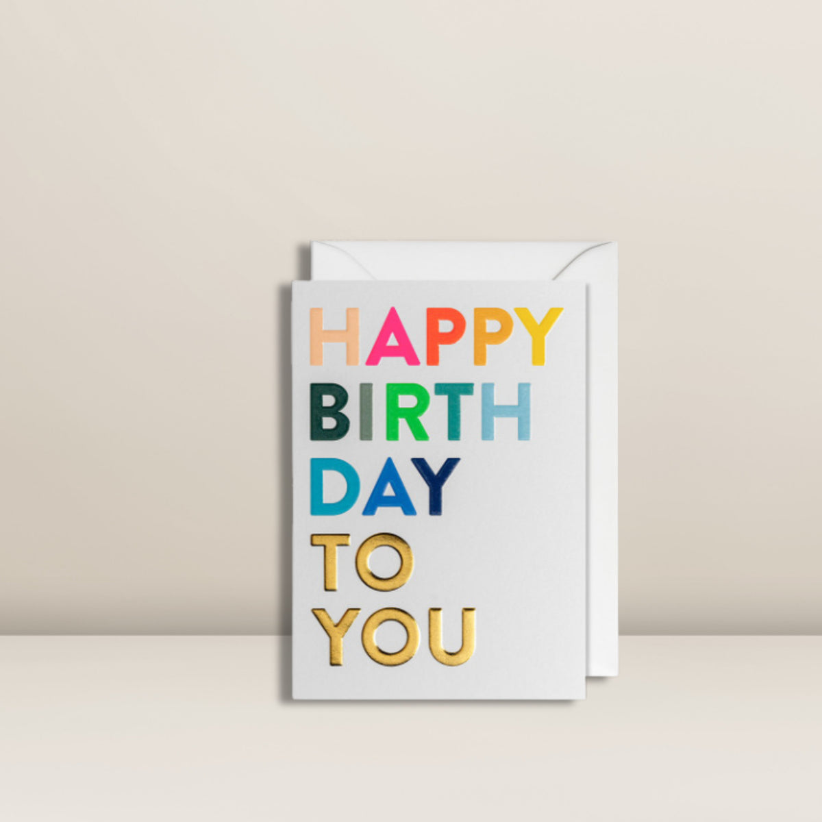 Die Grußkarte „Happy Birthday Bunt“ von Pacster besticht durch elegante, goldgeprägte und farbenfrohe Buchstaben, die den Schriftzug „HAPPY BIRTHDAY TO YOU“ zieren. Sie steht aufrecht vor dem passenden Umschlag auf einem beigen Untergrund. Entdecke hochwertige Geschenkideen bei Pacster.