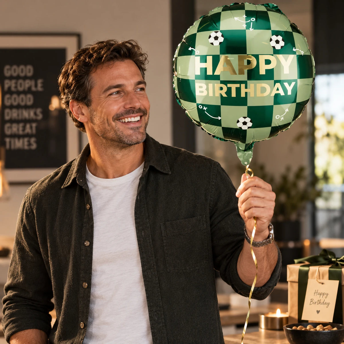 Ein lächelnder Mann in einem Freizeithemd hält den Pacster-Ballon „Happy Birthday Football“ in der Hand – einen grünen Ballon im Fußball-Design. Auf dem Tisch hinter ihm stehen Geschenktüten, ein Heliumballon und eine beleuchtete Geburtstagstorte, bereit, mit dem perfekten Geburtstagsgeschenk zu feiern. Entdecke hochwertige Geschenkideen bei Pacster.