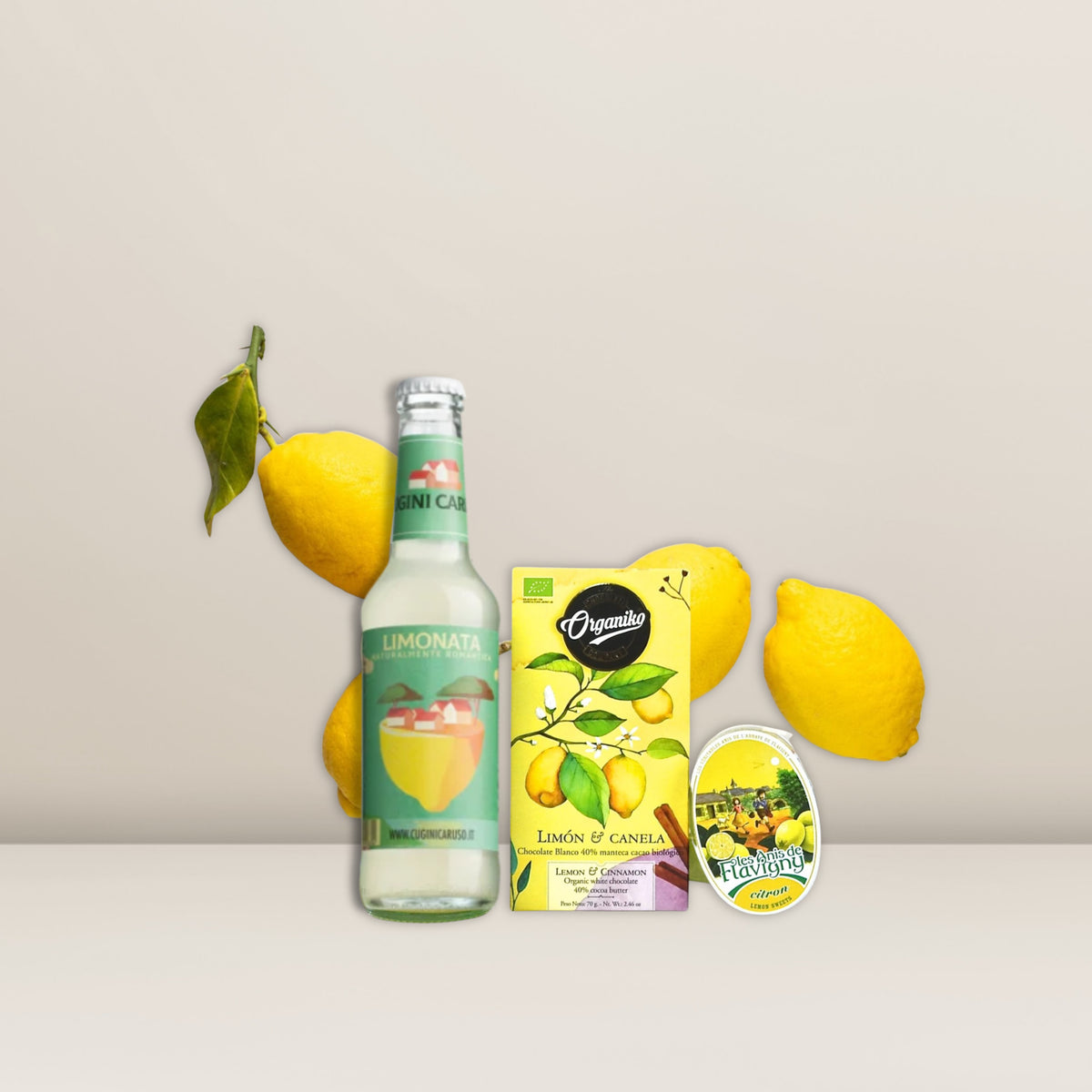 Ein kunstvoll zusammengestelltes „Pacster Sauer macht lustig“-Geschenkset enthält eine Flasche Bio-Zitronenlimonade, eine Schachtel Bio-Schokolade „Limón & Canela“-Tee, einen zitronenförmigen Behälter und drei frische Zitronen mit Blättern, alles auf einer hellen Unterlage vor einem blassen Hintergrund angerichtet – das perfekte Ensemble für eine elegante Geschenktüte. Entdecke hochwertige Geschenkideen bei Pacster.